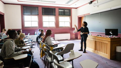 A female professor standing in the front of the classroom teaching to a room of students seated at their desks.