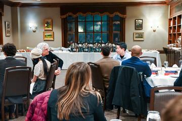 People seated at round tables in a dining room, conversing during a formal event with a buffet set up in the background.