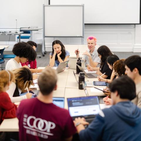 A group of students sits around a table in a classroom, collaborating while an instructor leads the discussion.
