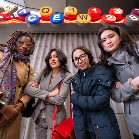 Four people stand together outdoors under colorful signage, posing confidently with arms crossed.