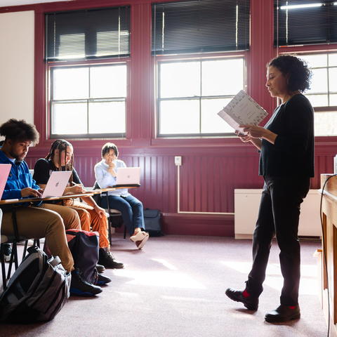 Instructor stands at the front of a classroom while students sit with laptops taking notes.