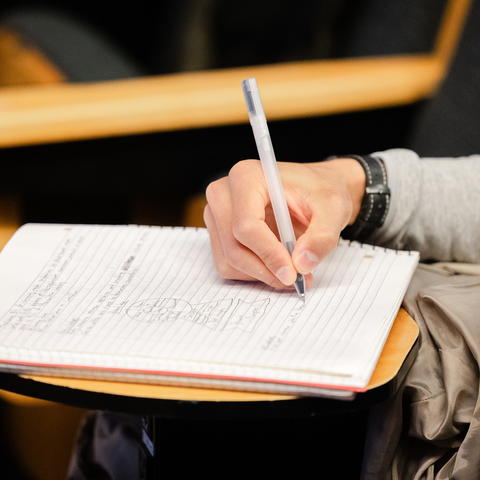 Person writes notes in a notebook while seated in a classroom.
