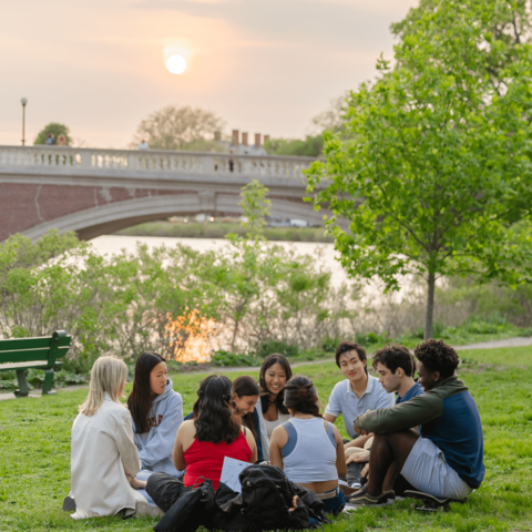 Group of students sitting in a circle on grass near a river at sunset.