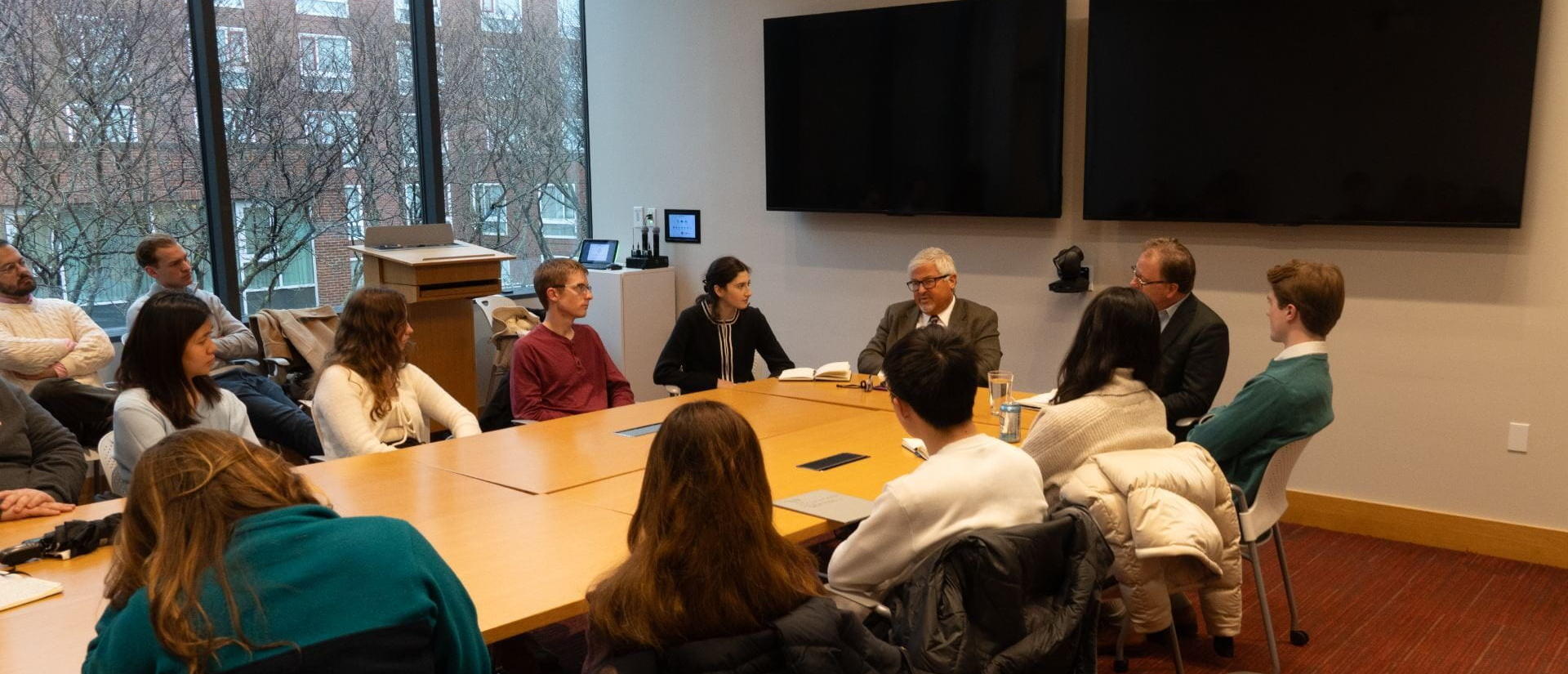 A room of students sitting around a table listening to a speaker.