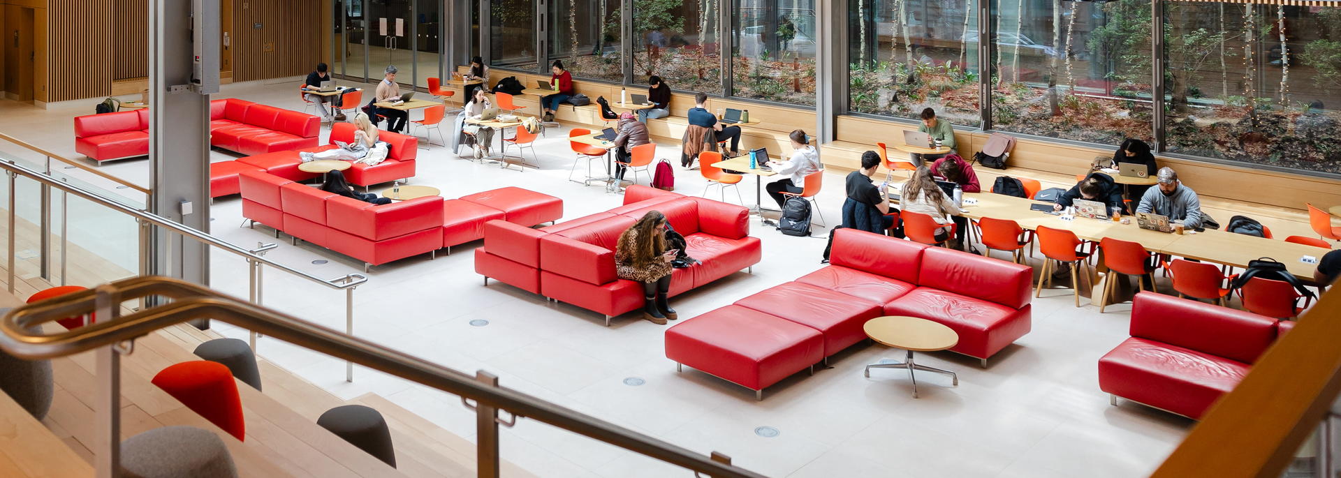 A bright, multi-level atrium with large windows where people sit, study, and socialize on red seating and tables.