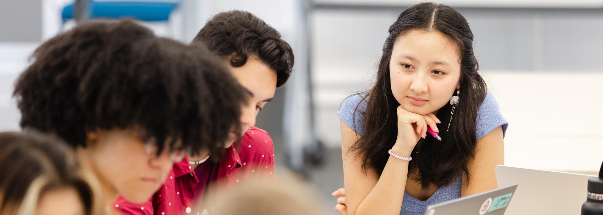 A group of students works together at a table, with one student thoughtfully looking at a laptop.
