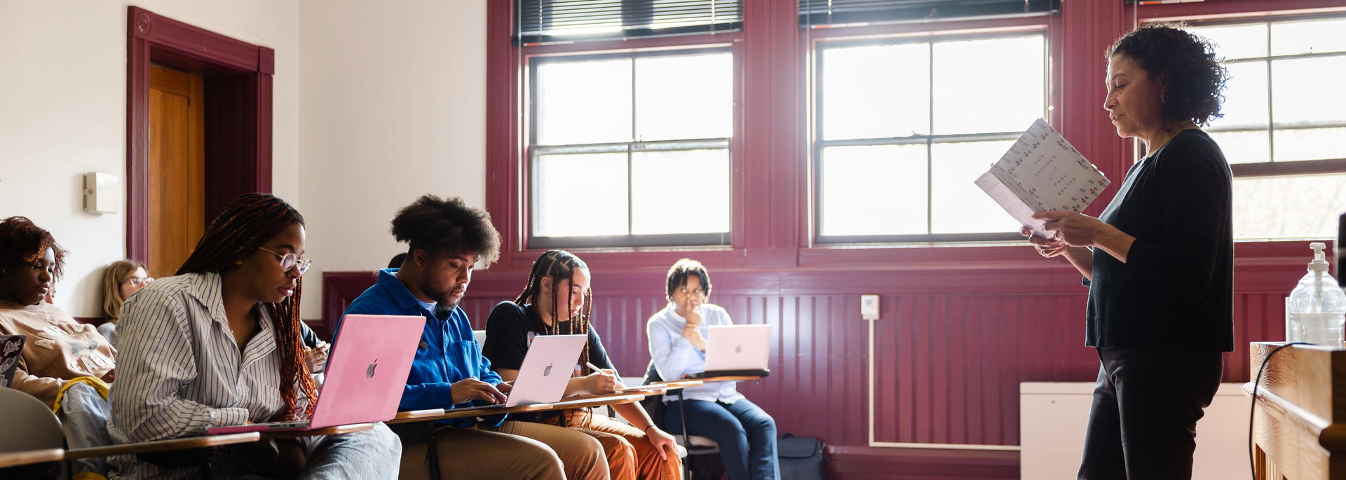 Instructor stands at the front of a classroom while students sit with laptops taking notes.