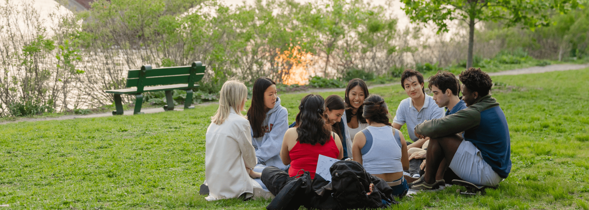 Group of students sitting in a circle on grass near a river at sunset.